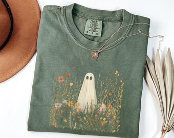 May include: Sage green t-shirt featuring a ghost graphic amidst wildflowers. The shirt has a crew neck and short sleeves. A gold necklace with a small pendant rests on the shirt. A brown hat is visible in the upper left corner.