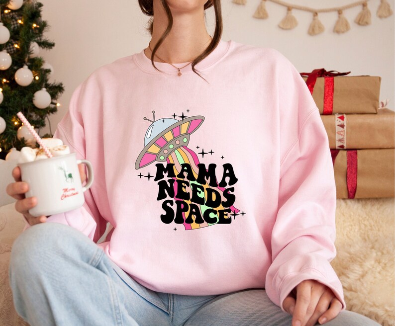 Puede incluir: Sudadera rosa con un gr&aacute;fico de un OVNI de colores arco&iacute;ris y el texto "Mama Needs Space" en negro.