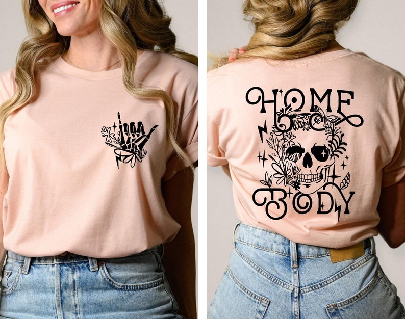 May include: A light pink t-shirt with a black skull and floral design on the back. The front of the shirt has a black hand with a floral design. The text on the back of the shirt reads "Home Body".