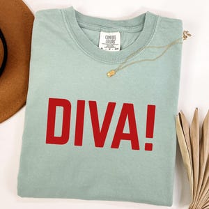 May include: A sage green short-sleeved t-shirt featuring the word DIVA! printed in bold red capital letters across the chest. The shirt has a crew neckline and a white interior neck label with the brand name Comfort Colors visible.