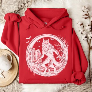 May include: A red hoodie featuring a white graphic of a werewolf standing in front of a haunted house, surrounded by a circular frame. Bats fly in the background. The hoodie has a drawstring hood and a kangaroo pocket.