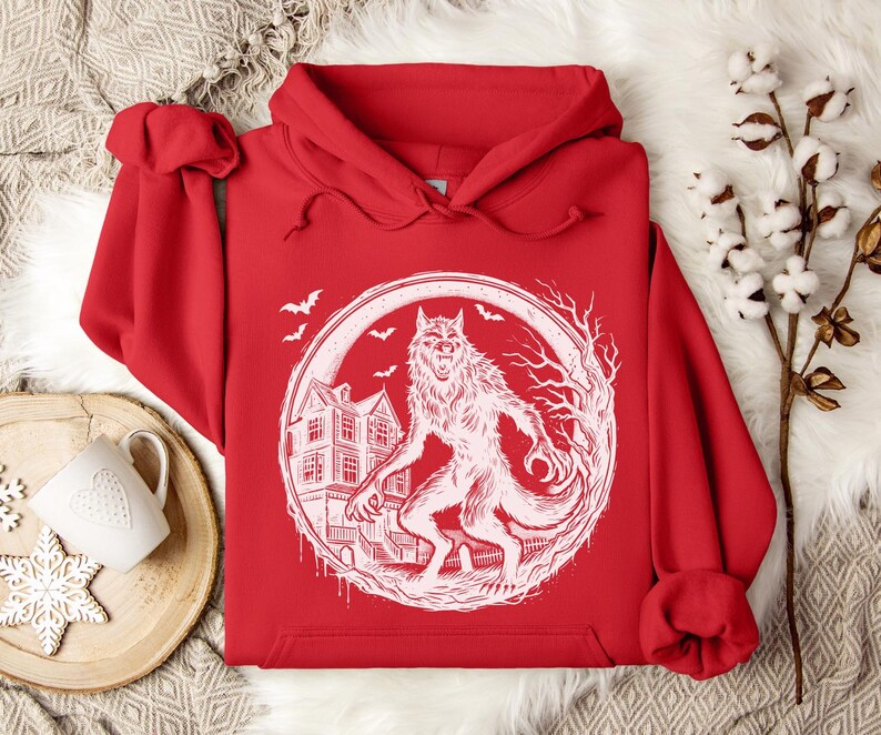 May include: A red hoodie featuring a white graphic of a werewolf standing in front of a haunted house, surrounded by a circular frame. Bats fly in the background. The hoodie has a drawstring hood and a kangaroo pocket.