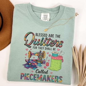 May include: A light green t-shirt with the text "Blessed are the Quilters for they shall be called Peacemakers." The shirt features a colorful sewing machine, spools of thread, and other quilting-related graphics.