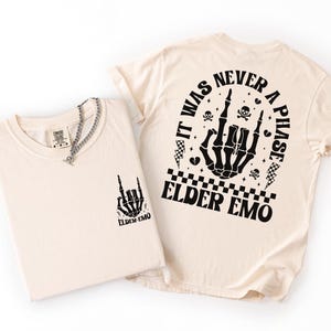 K&ouml;nnte beinhalten: Zwei cremefarbene T-Shirts mit schwarzen Grafikdesigns. Ein Shirt zeigt eine Skeletthand mit dem Text "ELDER EMO". Das andere Shirt hat ein gr&ouml;&szlig;eres Design auf dem R&uuml;cken mit dem Text "IT WAS NEVER A PHASE ELDER EMO". Eine silberne Halskette befindet sich auf einem Shirt.