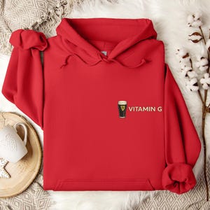 May include: A bright red hoodie with a drawstring hood. The hoodie features a small graphic of a pint of stout beer with the text "VITAMIN G" printed on the front. The hoodie is made of a soft material.