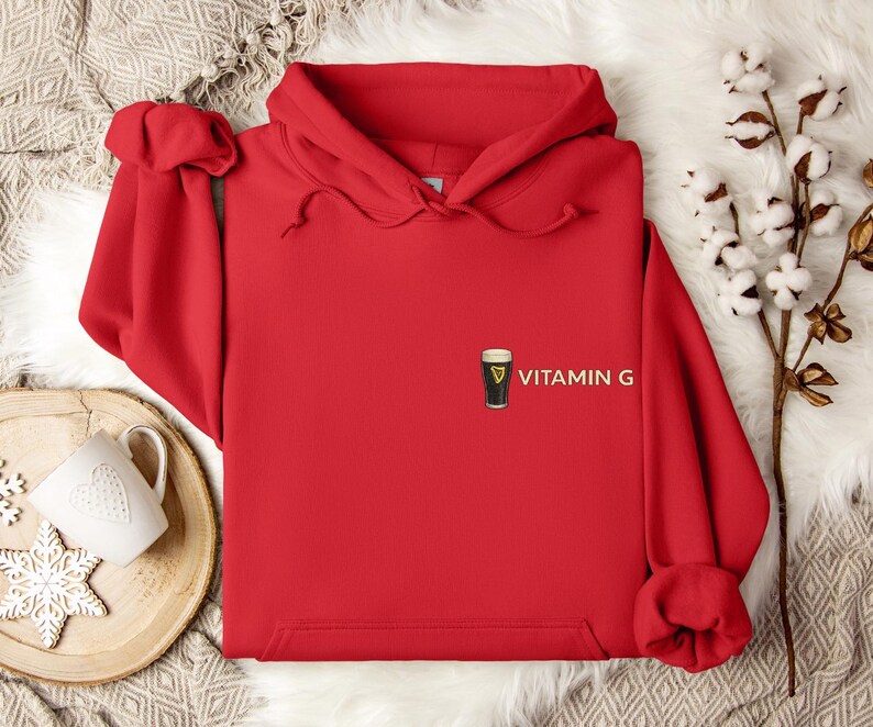 May include: A bright red hoodie with a drawstring hood. The hoodie features a small graphic of a pint of stout beer with the text "VITAMIN G" printed on the front. The hoodie is made of a soft material.