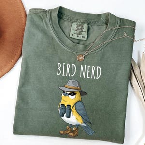 Op de afbeelding: Olijfgroen t-shirt met de tekst "BIRD NERD" en een cartoonvogel met een hoed, verrekijker en laarzen. Een gouden ketting ligt over het shirt. Het shirt ligt plat op een wit oppervlak.