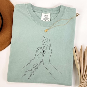May include: A sage green t-shirt with a black line drawing of a human hand and a dog paw touching. A gold necklace with a small pendant rests on the shirt. The shirt has a Comfort Colors tag.