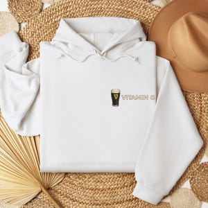 May include: A white hooded sweatshirt with a small graphic of a pint of stout beer and the text "VITAMIN G" in gold. The sweatshirt is displayed on a woven mat with a brown hat.