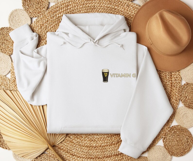 May include: A white hooded sweatshirt with a small graphic of a pint of stout beer and the text "VITAMIN G" in gold. The sweatshirt is displayed on a woven mat with a brown hat.