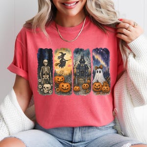 May include: Coral-colored t-shirt featuring four Halloween-themed illustrations. The designs include a skeleton, a witch on a broom, a haunted house, and a ghost with pumpkins. The artwork is in a watercolor style.
