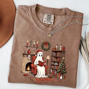 Op de afbeelding: Bruin Comfort Colors T-shirt met een cartoon spook dat een kerstmuts draagt en een boek leest bij een open haard. Het ontwerp bevat een kerstboom, een boekenkast en een krans. Het shirt heeft een ronde hals en korte mouwen.
