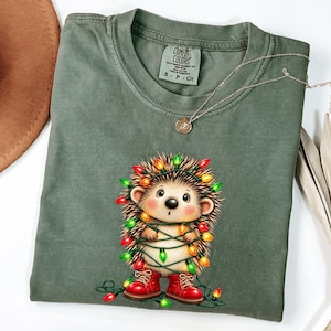 May include: An olive green t-shirt featuring a graphic of a small hedgehog wrapped in colorful Christmas string lights and wearing red boots. The shirt has a crew neckline and a Comfort Colors brand label printed on the inside collar.