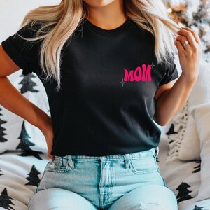 Puede incluir: Una camiseta negra con un gr&aacute;fico rosa de "MOM" en la parte delantera. La camiseta tiene un corte holgado y est&aacute; hecha de una tela suave y c&oacute;moda.