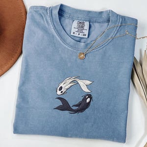 Puede incluir: Una camiseta azul de cuello redondo con un diseño bordado de dos peces koi, uno blanco y otro negro, dispuestos en un patrón circular de yin-yang. La camiseta tiene una etiqueta de la marca Comfort Colors en el cuello con la talla S-P-CH.