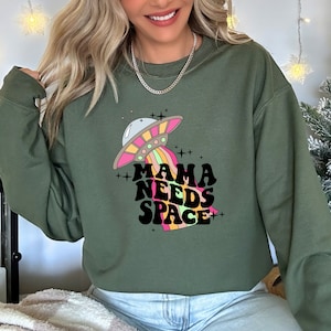 Puede incluir: Sudadera verde con un gr&aacute;fico de un platillo volante y el texto "Mama Needs Space" en una fuente retro.