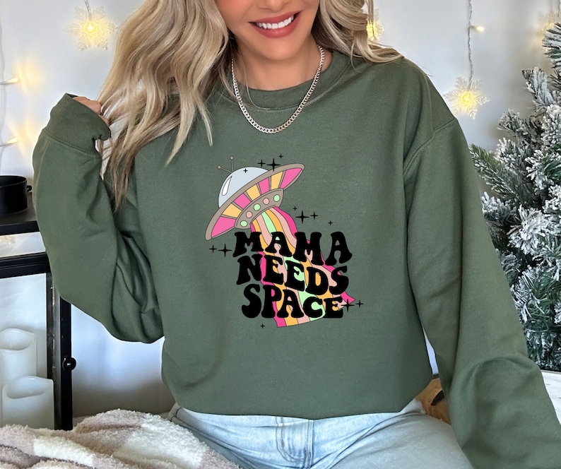 Puede incluir: Sudadera verde con un gr&aacute;fico de un platillo volante y el texto "Mama Needs Space" en una fuente retro.