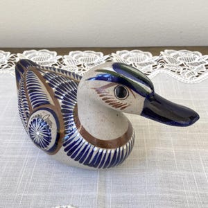 May include: Ceramic duck figurine with a detailed design. The duck features a white body with blue, brown, and green accents. The wings and tail have a blue and white pattern. The beak is dark blue.