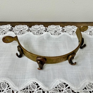 May include: A vintage brass decorative ring with three curved hooks and two rounded end pieces. The ring is set on a white lace tablecloth and a wooden surface.
