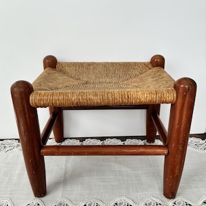 May include: A small wooden stool featuring four thick, rounded legs with a warm brown finish and a woven rush seat. The seat is constructed from natural tan fibers arranged in a square pattern, supported by a sturdy frame with horizontal stretchers.