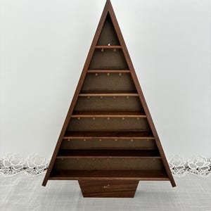 May include: A triangular, tree-shaped wooden display shelf with a dark brown finish. The unit features six horizontal shelves, each equipped with small gold-toned metal hooks along the underside for hanging items. The base is a solid wooden block.