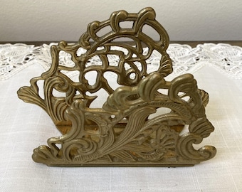 May include: Ornate brass napkin holder with a detailed, scrolling floral design. The vintage piece features a rectangular base and a curved, openwork top, showcasing a classic, elegant style. The brass has a warm, aged patina.