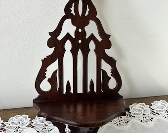 May include: A dark brown wooden corner shelf with an elaborate openwork design. The shelf has a curved front and a decorative back panel with cut-out shapes. The shelf is held by a hand against a white wall.