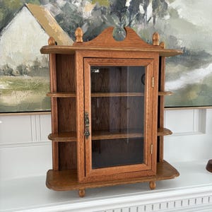 May include: A medium-brown wooden wall cabinet featuring a central glass-paneled door with a decorative metal handle. The unit includes three open shelves on each side, a flat top with three small finials, and a base supported by four small feet.
