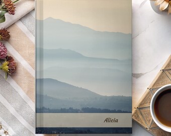 May include: A hardcover journal featuring a landscape image of layered blue mountains under a pale yellow sky. The name "Alicia" is printed in a dark font at the bottom. The journal is surrounded by flowers and a cup of coffee.