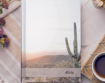 Op de afbeelding: Een hardcover notitieboek met een woestijnlandschap, een saguaro cactus en de naam "Alicia" op de kaft. Het notitieboek heeft een witte rug en een foto van een zonsondergang in de woestijn.