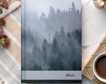 May include: A hardcover journal featuring a misty forest scene. The cover displays a gray and white band with the name "Alicia" in a script font. The journal is on a striped beige and white cloth.