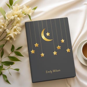 Op de afbeelding: Een donkergrijs notitieboek met een ontwerp van een gouden halve maan en sterren. Het notitieboek heeft de naam "Emily Wilson" op de voorkant.