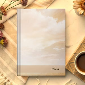May include: A hardcover journal with a soft, cloud-like design in shades of beige and cream. The name "Alicia" is printed in a delicate script at the bottom. The journal is placed on a cream-colored surface with flowers and a cup of coffee.
