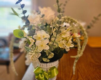 May include: A white ceramic vase with a green ribbon, filled with white flowers and green foliage.