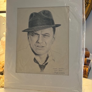 Peut inclure: Portrait au crayon de Frank Sinatra portant un fedora et une cravate. L'œuvre est dans un passe-partout blanc et scellée dans du plastique. Le texte indique "Frank Sinatra, Tony Rome, Joseph G. Forte".