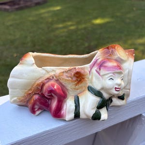 May include: A vintage ceramic planter in the shape of a smiling elf. The planter is cream-colored with accents of red, green, and brown. The elf is wearing a hat and a scarf. The planter is designed to hold small plants or flowers.