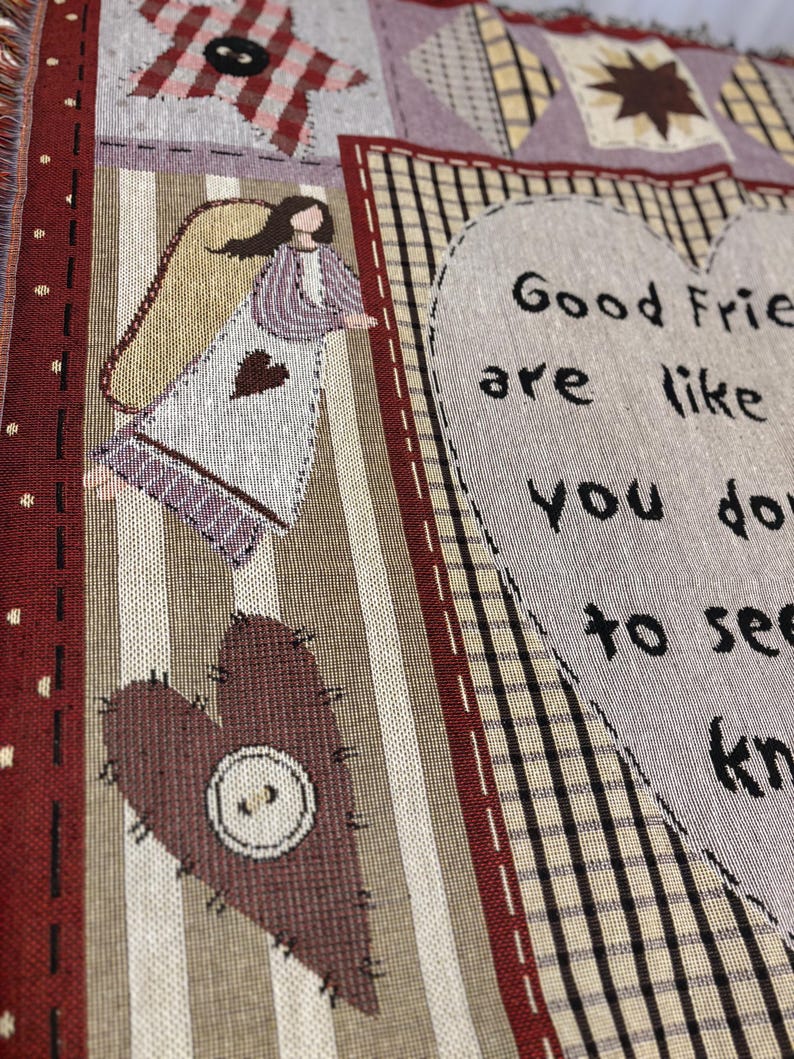 May include: A woven throw blanket with a patchwork design. The blanket features a heart with a button, an angel, and the text "Good Friends are like you don't have to see..." in black lettering. The color palette includes burgundy, beige, and purple.