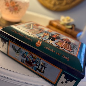 Könnte beinhalten: Eine grüne und blaue Vintage-Metallbox mit Scharnierdeckel. Die Box zeigt farbenfrohe Illustrationen von Menschen in traditioneller Kleidung und den Text "Lebkuchen und Honig". Die Box ist mit "E. Otto Schmidt Nürnberg" beschriftet.