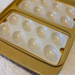 Pu&ograve; includere: Un contenitore per uova vintage in plastica gialla con due sezioni, ciascuna con una dozzina di rientranze a forma di uovo. L'interno &egrave; bianco e il contenitore &egrave; progettato per contenere le uova in modo sicuro. Il contenitore &egrave; su una superficie bianca.