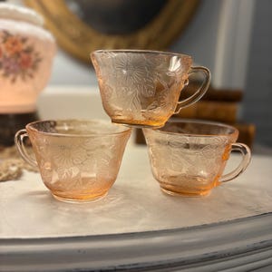 Puede incluir: Una taza de cerámica blanca con un asa negra y el texto "But First, Coffee" en negrita negra. La taza se encuentra sobre un fondo neutro, resaltando su diseño simple.