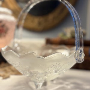 May include: A clear glass basket with a handle. The basket has a scalloped edge and is supported by three small feet. The glass has a frosted appearance with a floral design. The handle is arched and attached to the basket.