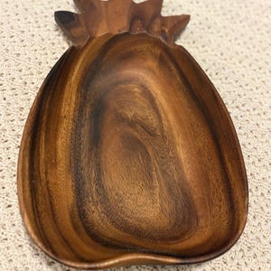 Puede incluir: Un cuenco de madera tallada con forma de piña, que presenta un color marrón intenso con patrones de veta de madera natural visibles. El cuenco tiene un interior cóncavo y liso y una sección superior de hojas distintiva, mostrando la textura orgánica.