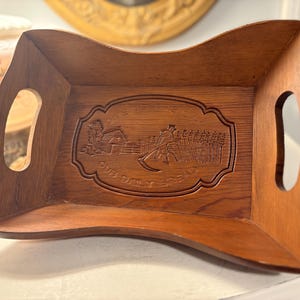May include: A wooden serving tray with a curved shape and two handle cutouts. The tray features a carved design with the words "Give us this day our daily bread" and a rural scene. The wood has a warm, brown tone.