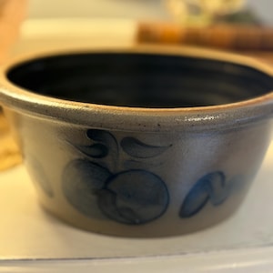 May include: A vintage stoneware bowl with a light beige exterior and a dark blue interior. The bowl features hand-painted blue fruit and leaf designs. The rim is slightly flared, and the bowl has a rustic, aged appearance.