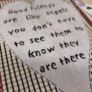 May include: A quilted fabric with the text "Good Friends are like angels, you don't have to see them to know they are there" in a heart shape. The background has a plaid pattern in shades of brown and cream.