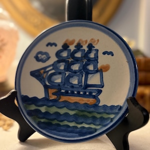 May include: A round ceramic plate with a hand-painted sailboat design. The plate features a blue and white color scheme with orange accents. The sailboat is depicted sailing on blue waves, with clouds and birds in the sky. The plate is displayed on a black stand.