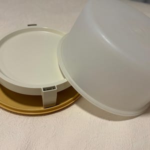 Peut inclure: Un récipient de stockage alimentaire en plastique en trois parties, blanc cassé et jaune. Le récipient comprend une base ronde, un plateau blanc avec des pieds et un couvercle blanc translucide. La base a deux poignées.