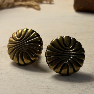 Puede incluir: Dos tiradores de armario de latón antiguo con un diseño detallado y arremolinado. Los tiradores tienen un acabado cálido y envejecido y están colocados sobre una superficie de madera blanca. Los tiradores son redondos y tienen una superficie decorativa y texturizada.