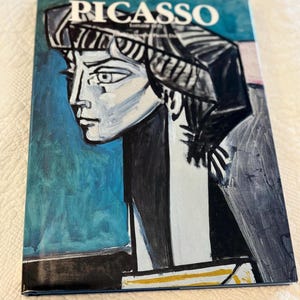Könnte beinhalten: Ein gebundenes Buch mit dem Titel PICASSO, das auf dem Einband ein stilisiertes Porträt einer Person im Profil zeigt. Das Kunstwerk verwendet kräftige schwarze, weiße und blaue Pinselstriche, um die Person mit einer dunklen Kopfbedeckung darzustellen.