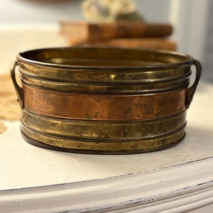 May include: A round metal basin featuring a wide central band of polished copper flanked by two brass bands with horizontal ridges. The vessel includes two small, curved metal handles attached to opposite sides of the exterior rim.